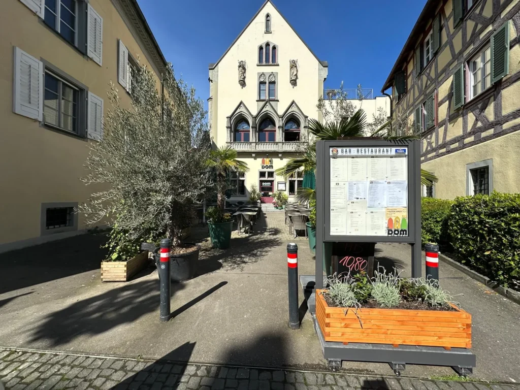 Weitwinkelblick auf den Innenhof und die gotische Fassade des The Dom Restaurant Konstanz mit Speisekarten-Vitrine und Olivenbäumen