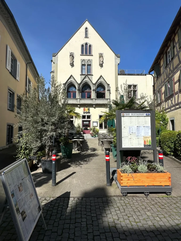 Sonniger Innenhof des The Dom in Konstanz mit Außenmenükarte, Olivenbäumen und gotischer Fassade