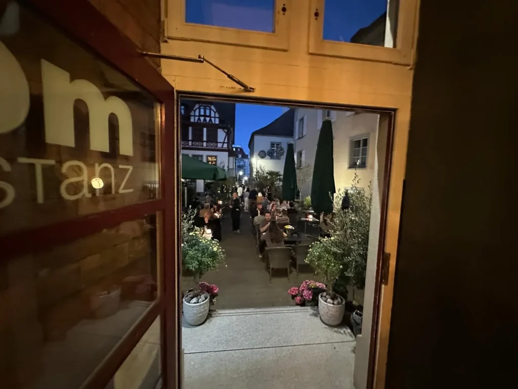 Blick durch die geoeffnete Eingangstuer von The Dom Konstanz auf die beleuchtete Abendterrasse