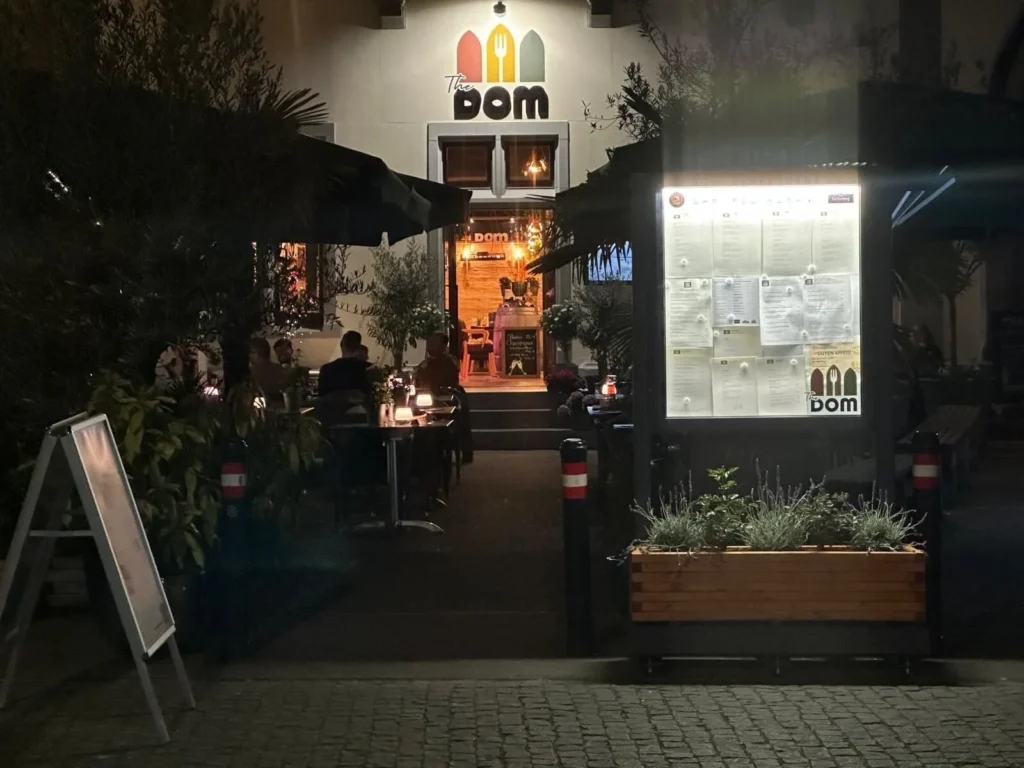 Beleuchteter Eingangsbereich von The Dom Konstanz bei Nacht mit Logo, Speisekarte und Terrasse
