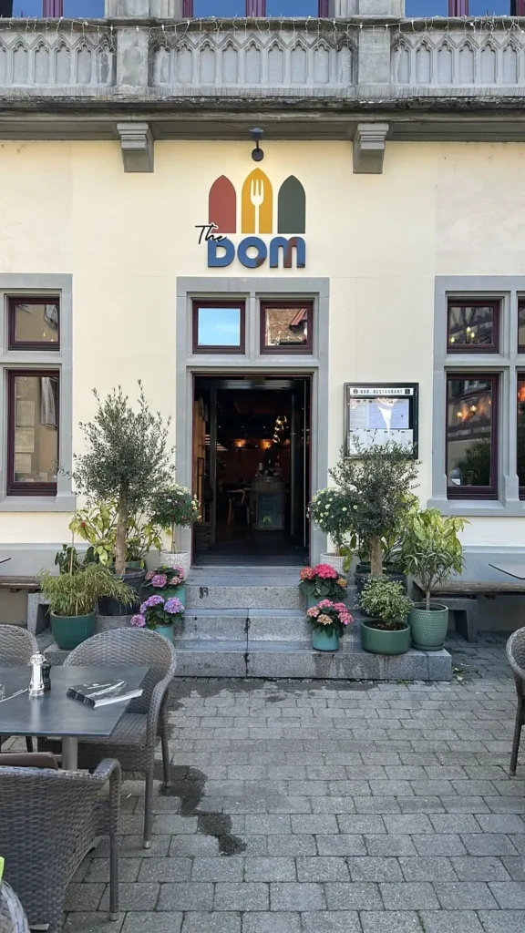 Eingang von The Dom Konstanz mit Logo, Olivenbaeumen und Blumen vor historischer Fassade