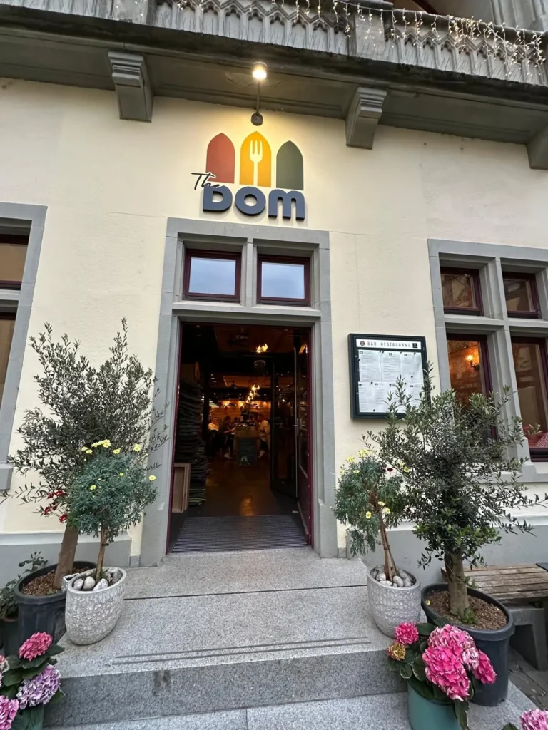Fassade des Restaurants The Dom Konstanz mit beleuchtetem Logo und Olivenbaeumen