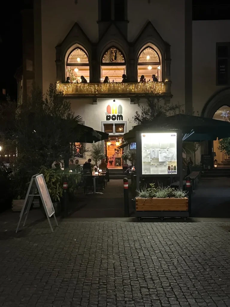 Beleuchtete gotische Fassade von The Dom Konstanz bei Nacht mit Gaesten auf dem Balkon und Speisekarte