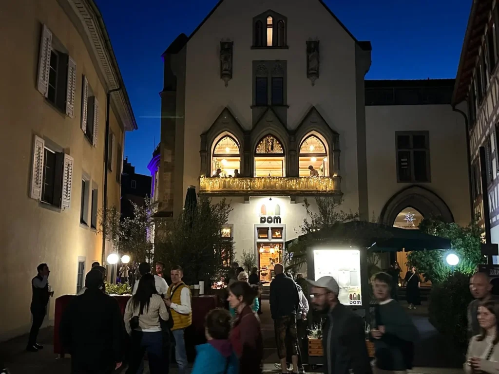 Beleuchtete gotische Fassade von The Dom Konstanz bei Nacht mit Lichterketten am Balkon und Gaesten