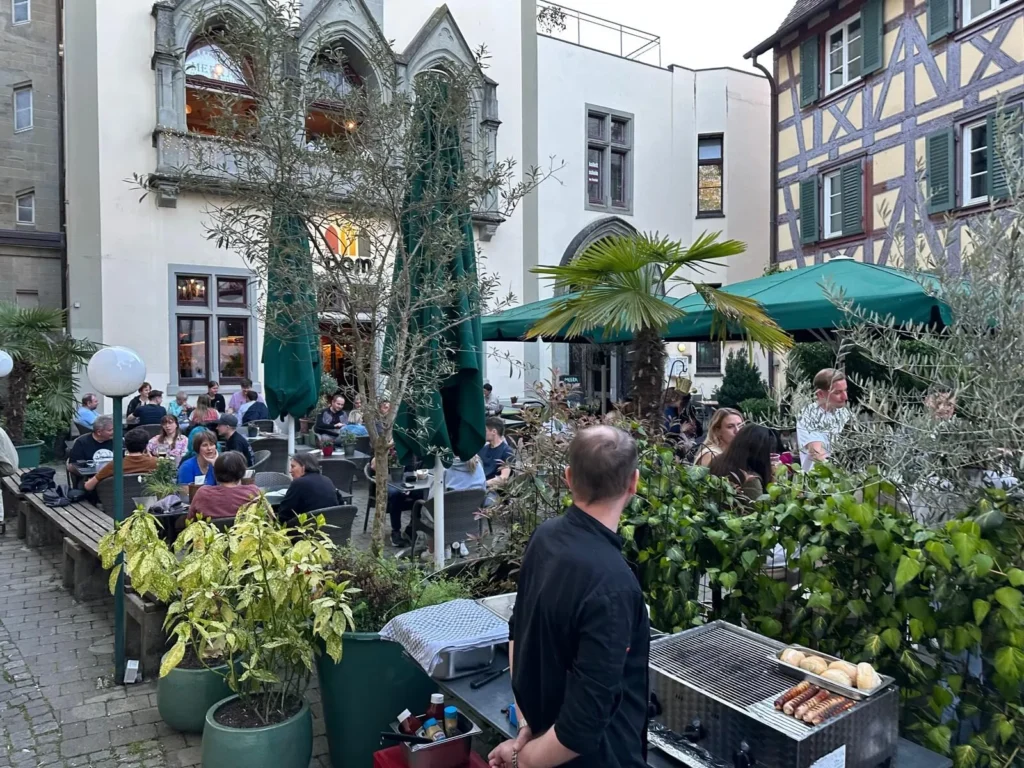 Koch am Grill im belebten Innenhof von The Dom Konstanz mit Olivenbaeumen und Fachwerkhaeusern
