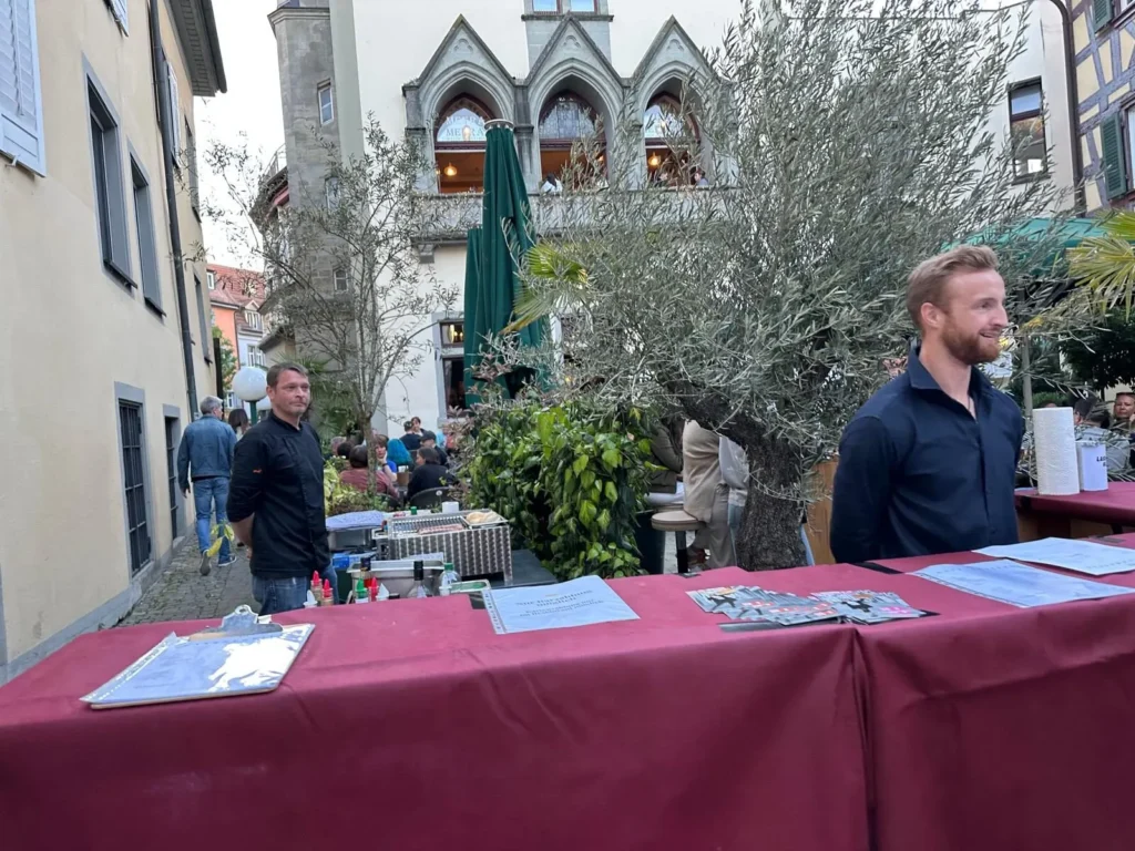 Zwei Mitarbeiter von The Dom Konstanz am Empfangstisch vor dem Restaurant mit Olivenbaum