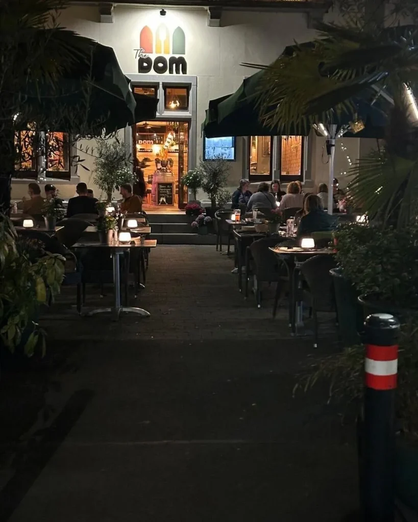 Belebte Terrasse von The Dom Konstanz am Abend mit Palmen, Gaesten und warmem Licht