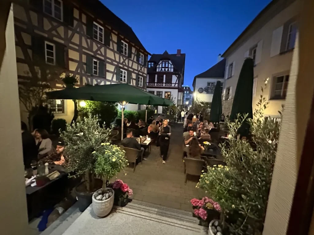 Bedienung auf der belebten Abendterrasse von The Dom Konstanz zwischen Fachwerkhaeusern und Olivenbaeumen